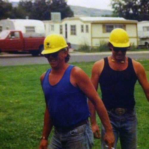 Two workers wearing yellow hard hats and tank tops walk across a grassy area, with a red pickup truck and mobile homes in the background.